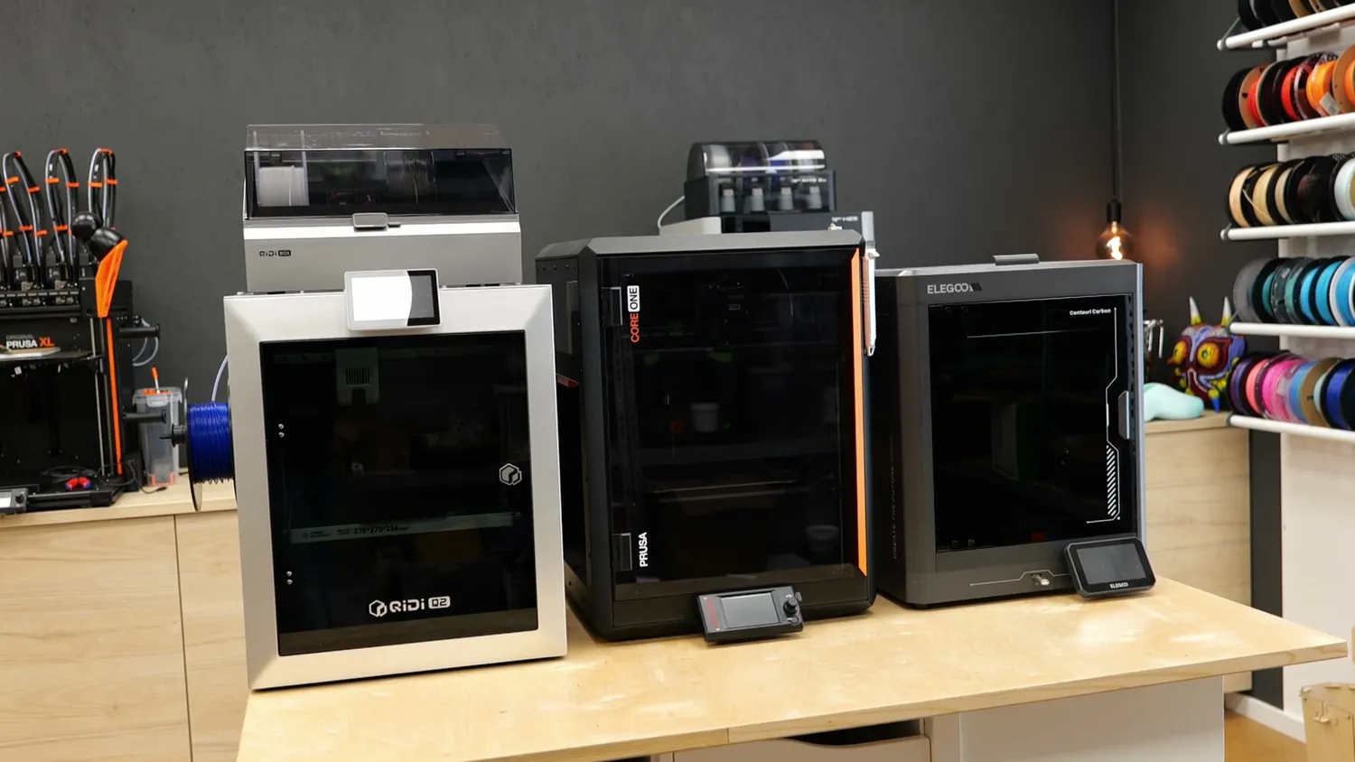 A QIDI Q2, a Prusa CORE One and an Elegoo Centauri Carbon standing side by side on a bench