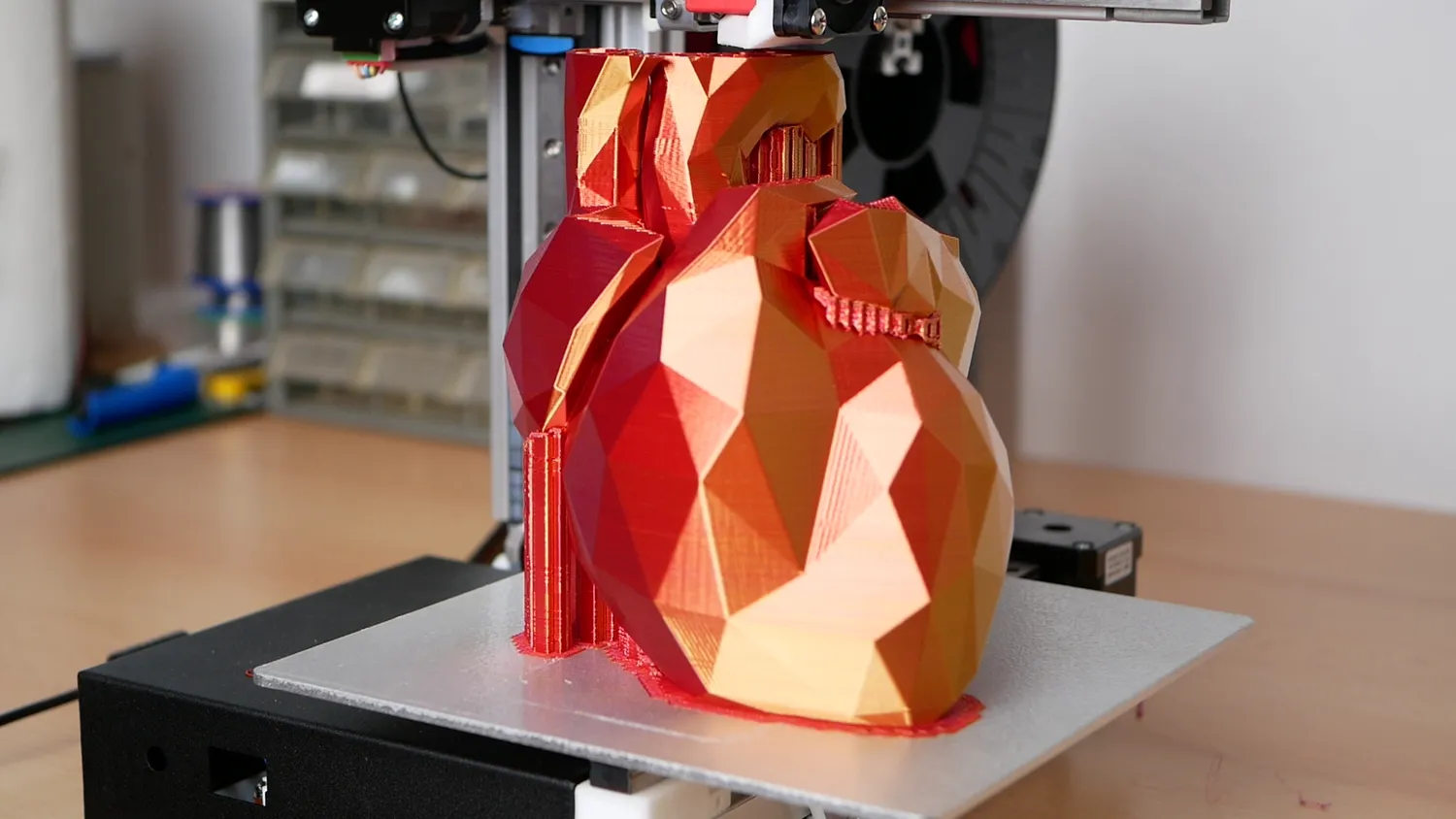 A large low-poly bunny in red-to-gold gradient filament, printing with a support tower alongside