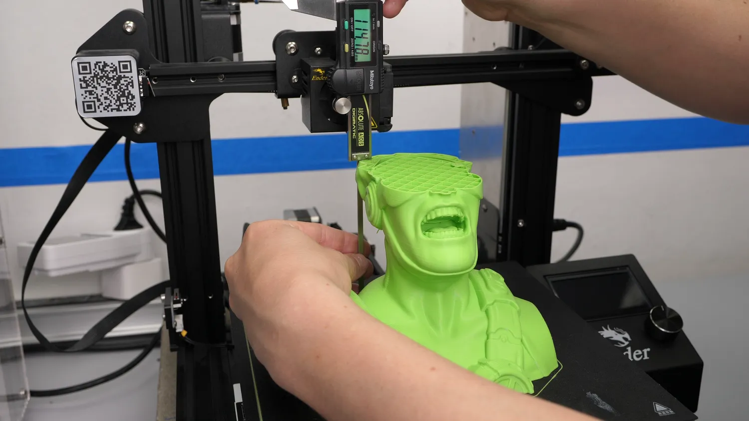 Digital calipers measuring the height of the failed green bust, held perpendicular and reaching all the way down to the bed