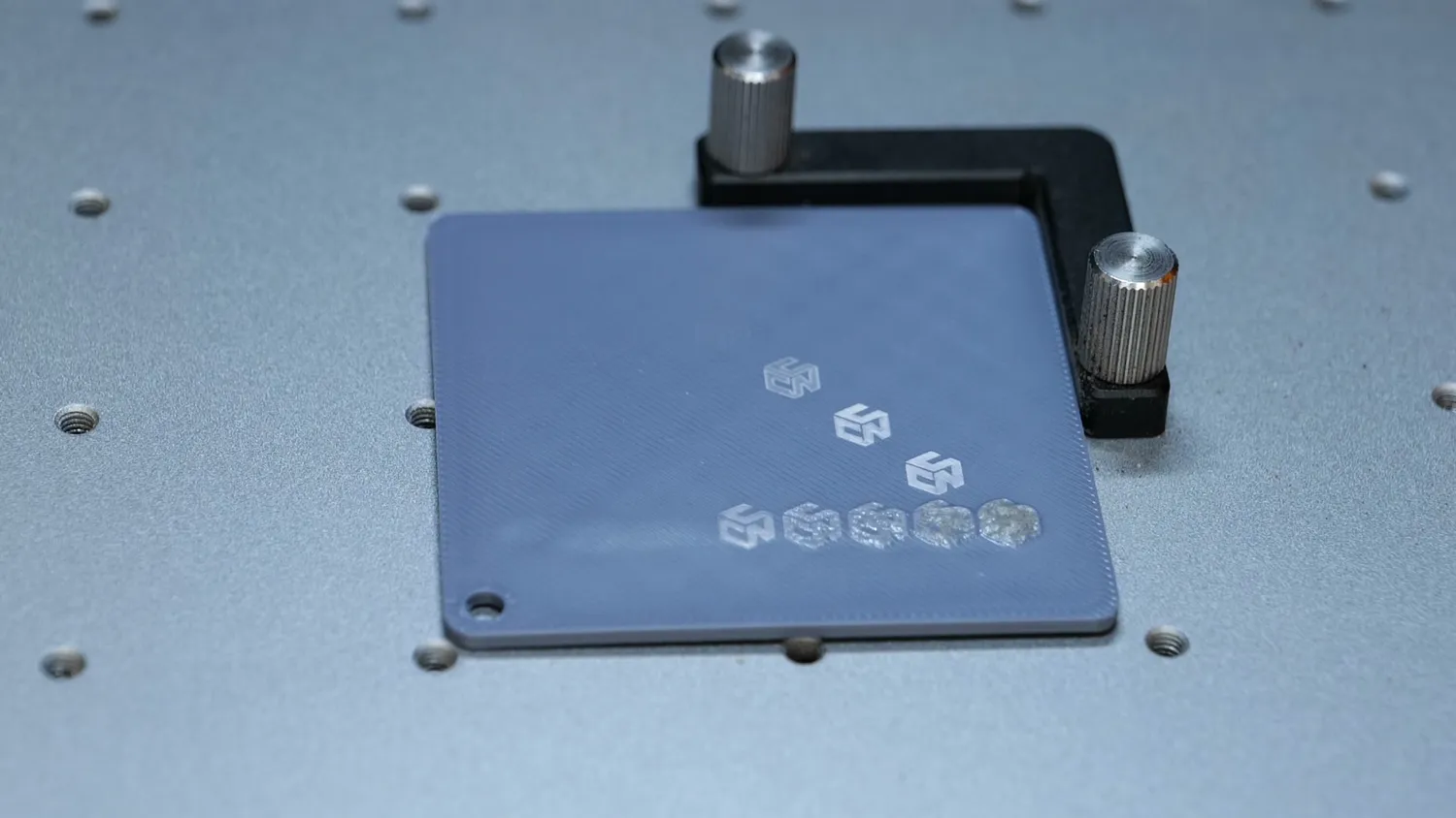 Grey 3D printed test plate on the laser bed with a row of marked CNC Kitchen logos