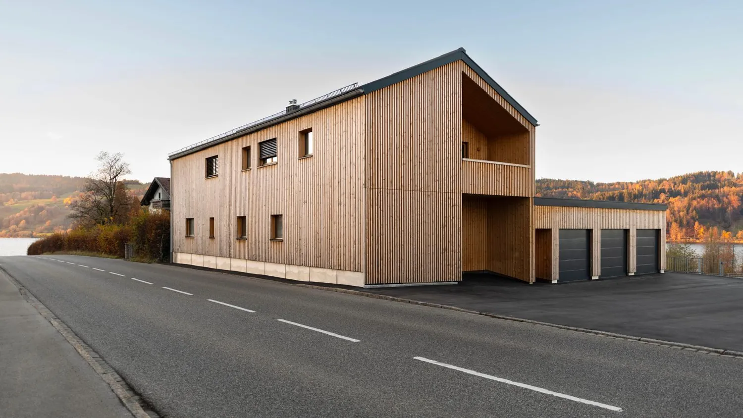 The CNC Kitchen warehouse in the Allgäu: a timber-clad former fire station with three roller doors, overlooking a lake and autumn hills